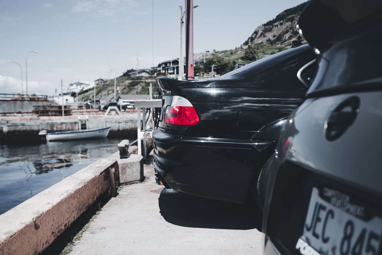 Close-up of classic black cars parked by a peaceful waterfront harbor on a sunny day.